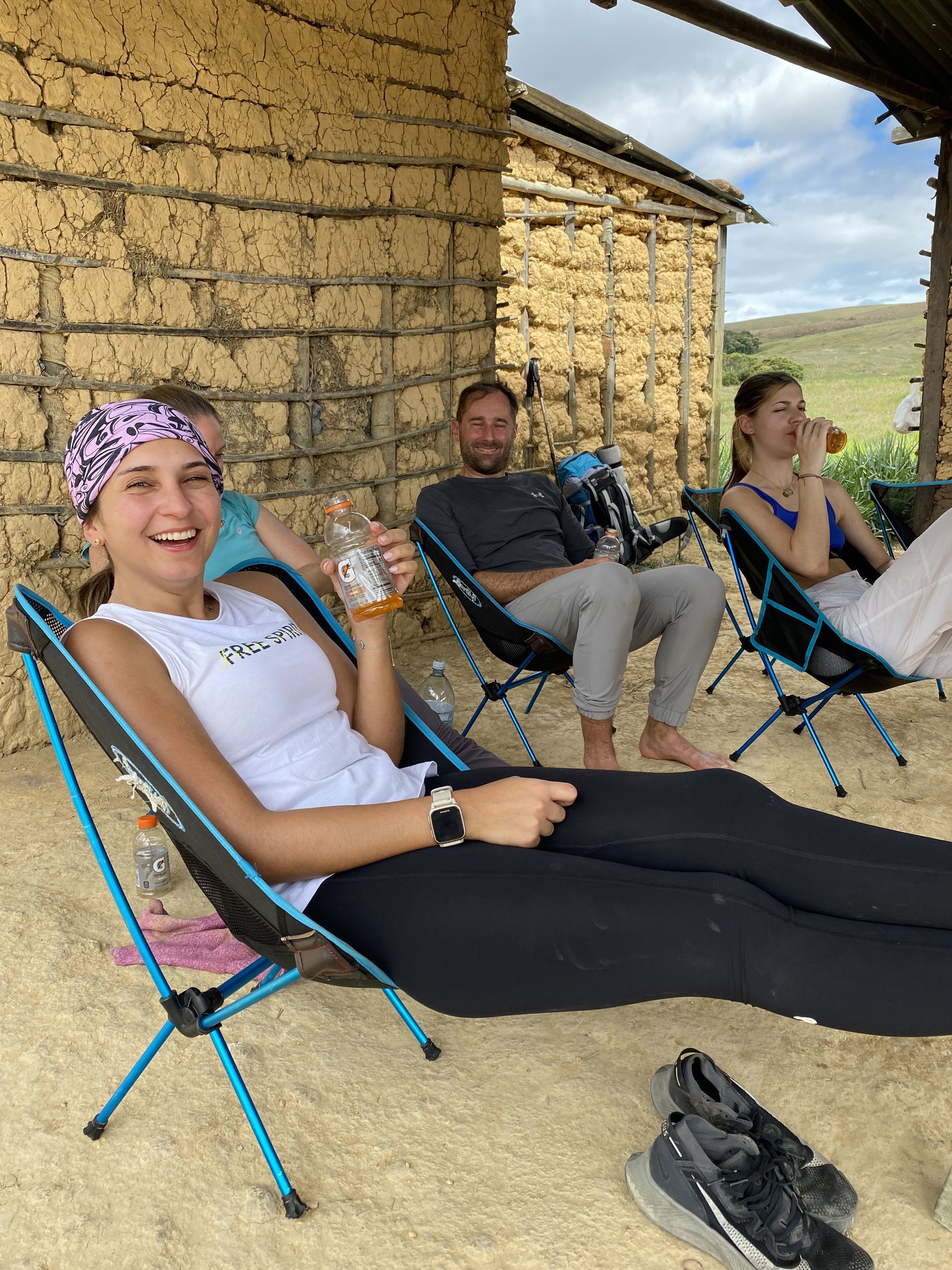 Three people relax in camping chairs under a rustic shelter. A woman smiles, holding a drink, while two others sit nearby, also with drinks.