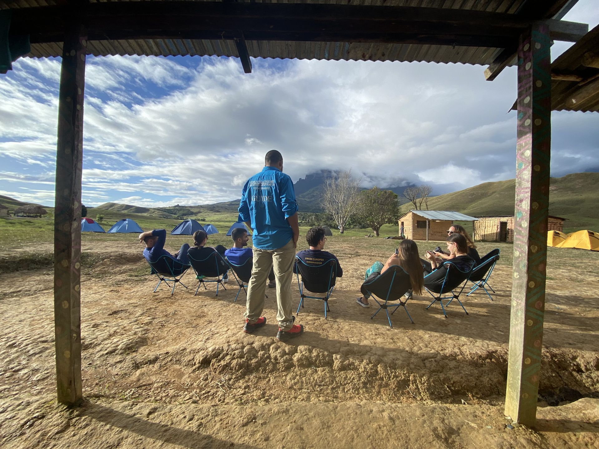 A group of people relaxing in camp chairs outdoors. One person stands facing them under a shelter. Cloudy sky and tents in the background.