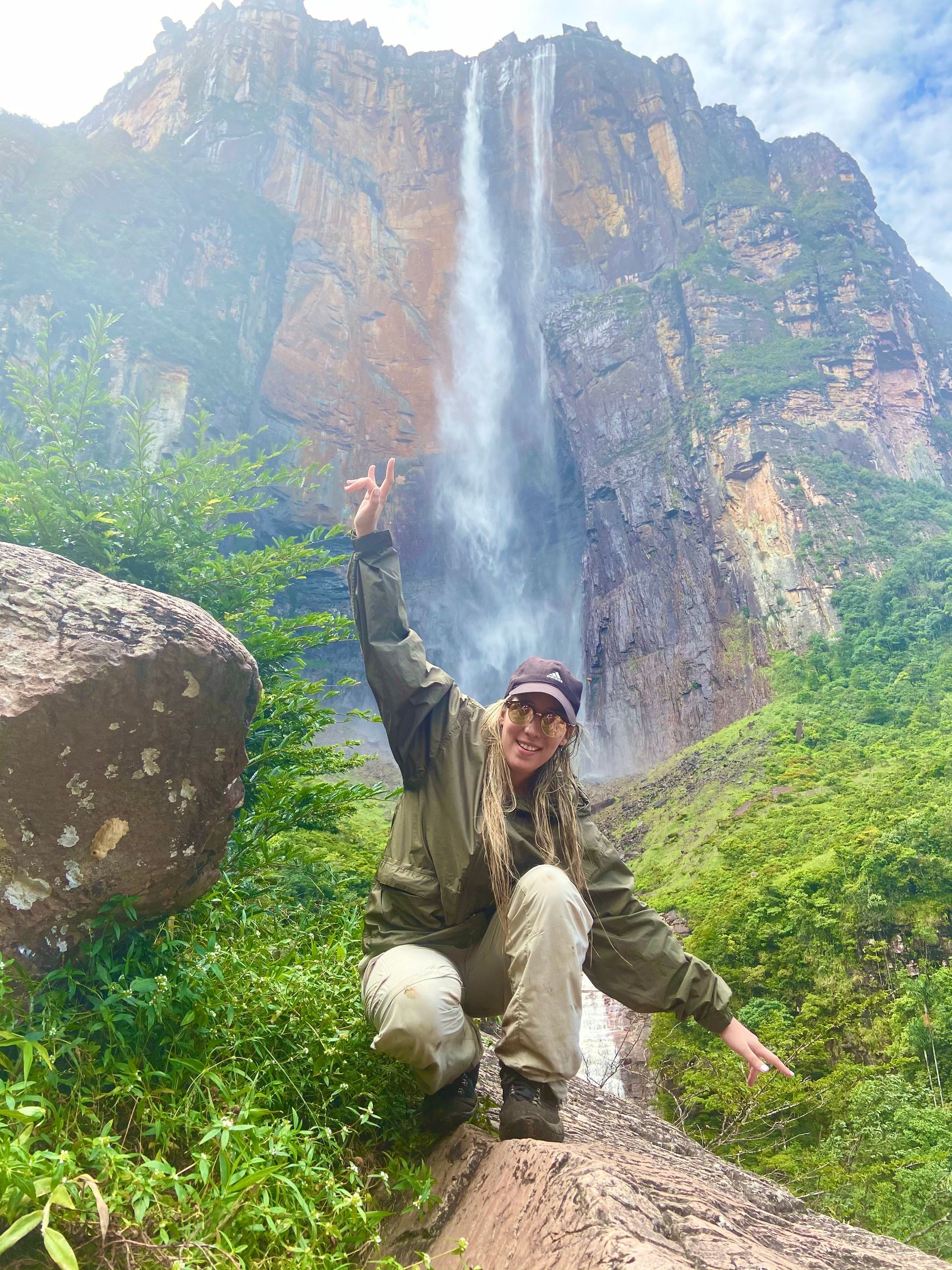 Woman kneels in front of Angel Falls, raising her arm in a peace sign. She wears olive green clothing and a baseball cap in a lush, green environment.