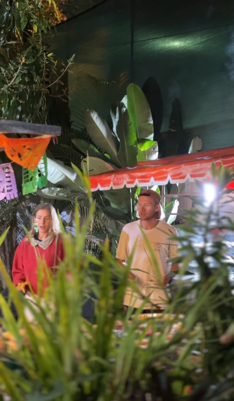 Two people stand at a food stall with red awning and tropical plants. The woman wears red; the man, a beige shirt.