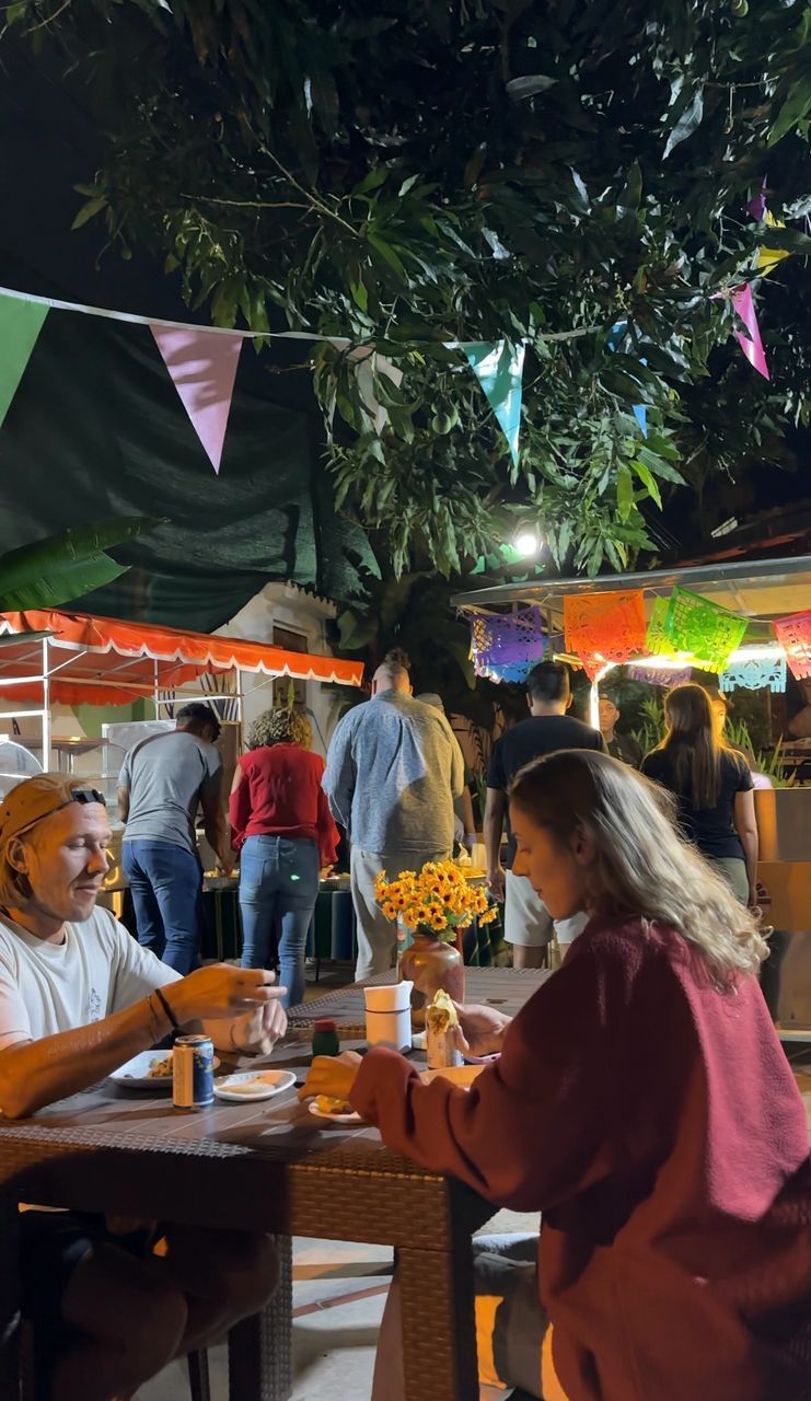 People at an outdoor night market, some eating at tables. Colorful bunting, food stalls, and a tree are in the background.