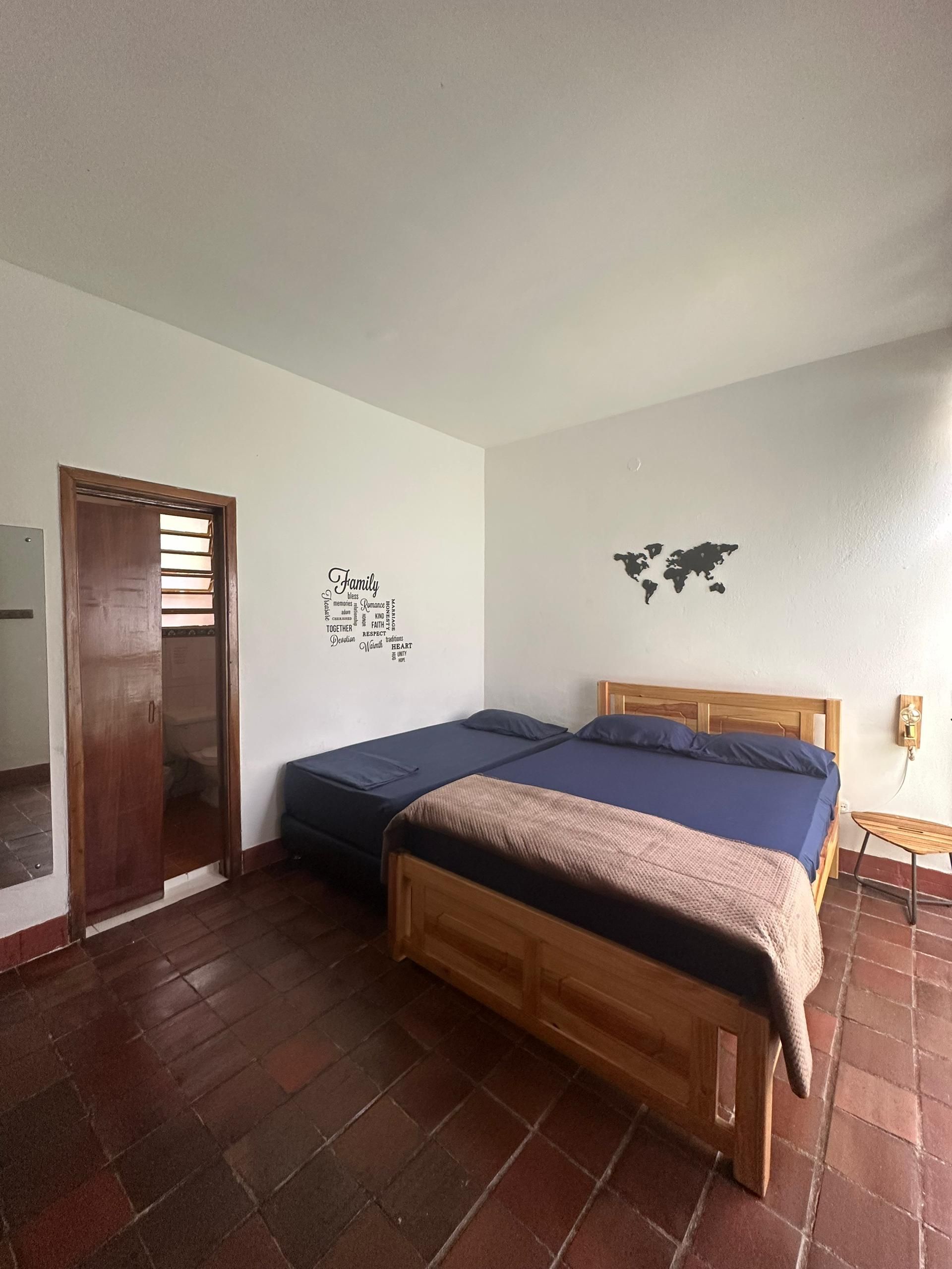 A bedroom with two twin beds with blue sheets and a brown blanket, brown tiled floor, and a wooden door.