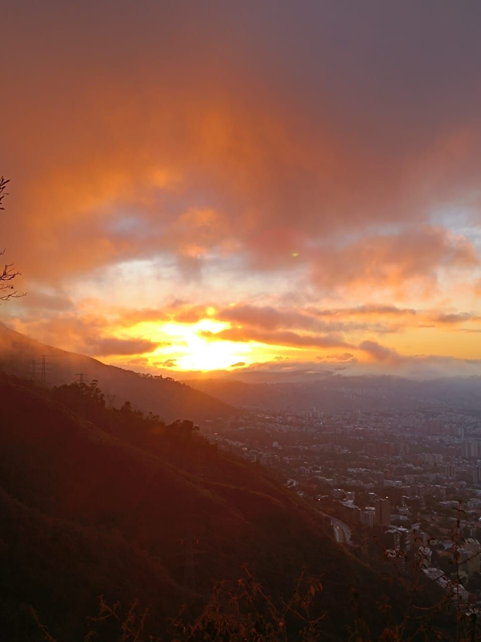 Sunset over a cityscape, viewed from a mountainside. Fiery orange and yellow sky; dark silhouette of a mountain in the foreground.
