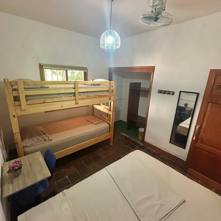A room with a bunk bed, a double bed, a small table, and a closet. A ceiling fan and a mirror are also visible.