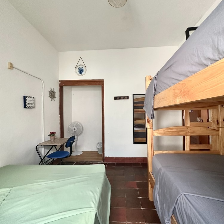 A hostel room with bunk beds and a single bed. A desk, fan, and open doorway are also visible.