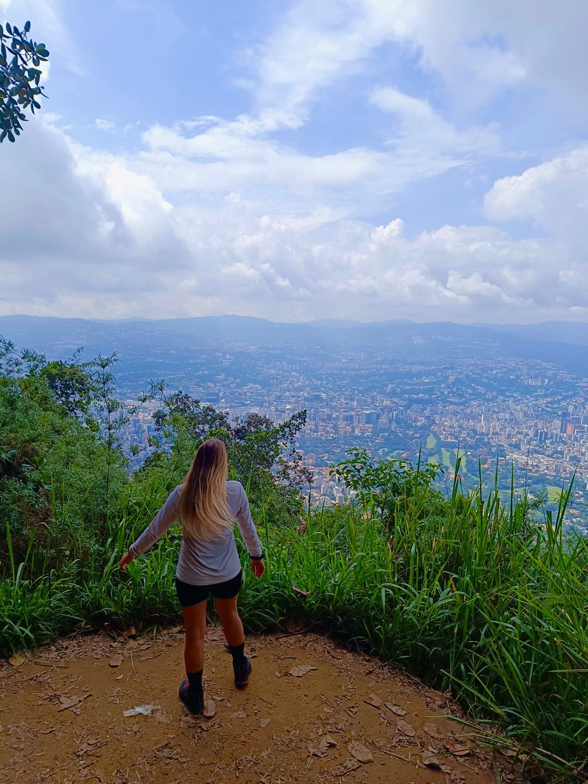 Woman with blonde hair looking out over a city from a green, hilly viewpoint under a cloudy sky.
