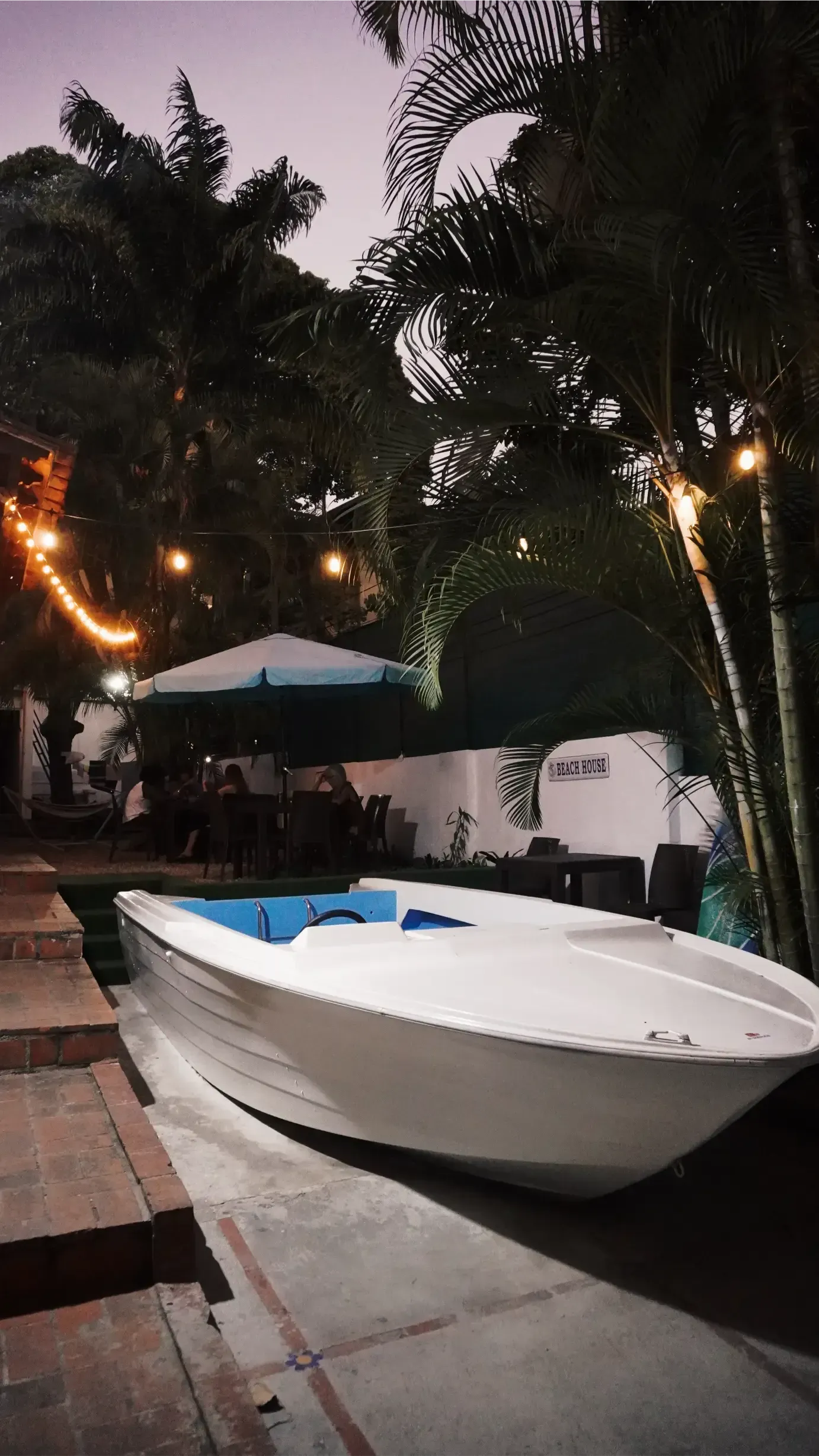 White boat on the ground near a patio with tables, string lights, and palm trees. Nighttime scene.