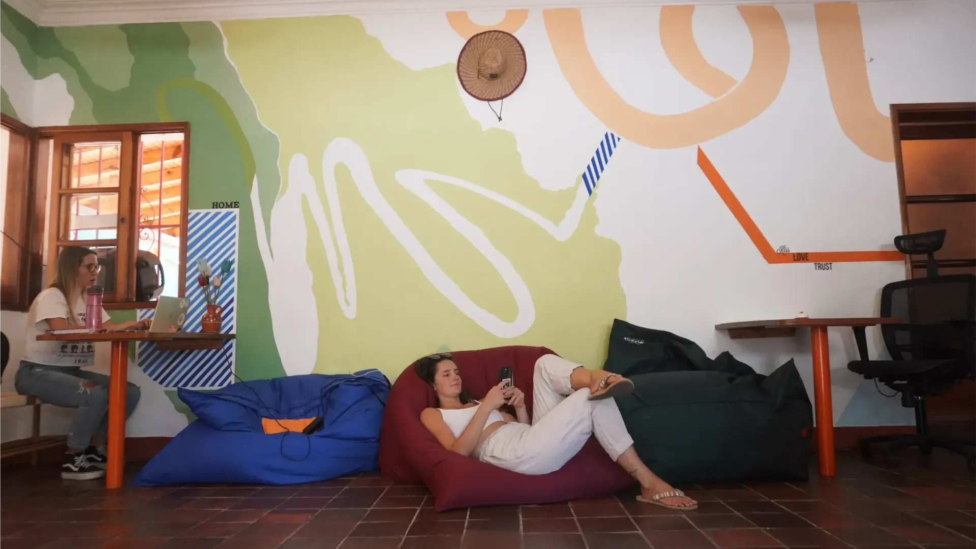 A woman relaxes in a red beanbag chair, looking at her phone in a brightly decorated room. Another person sits at a desk in the background.