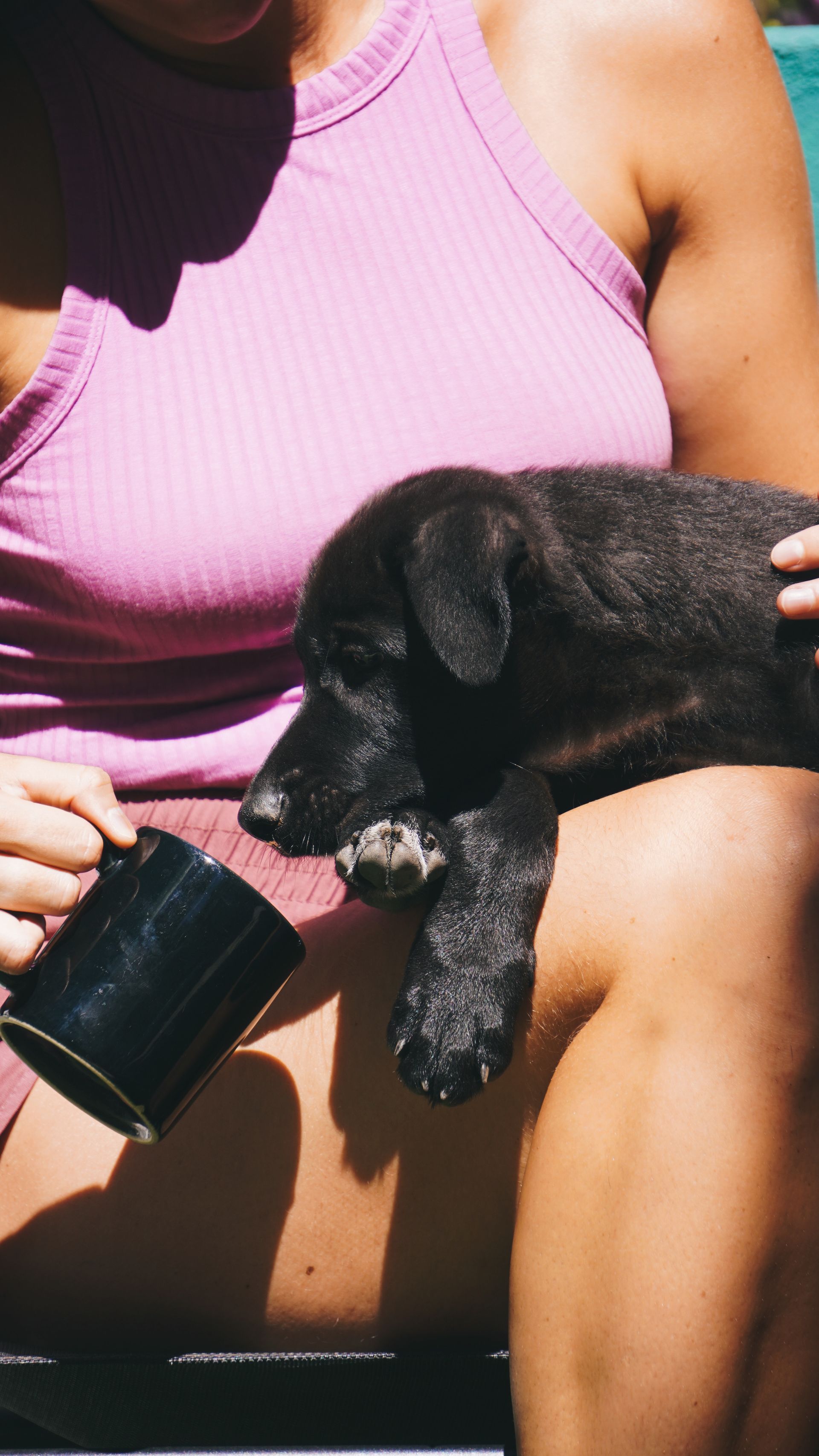 Woman in pink tank top holding a black puppy on her lap, with a black mug in her hand.