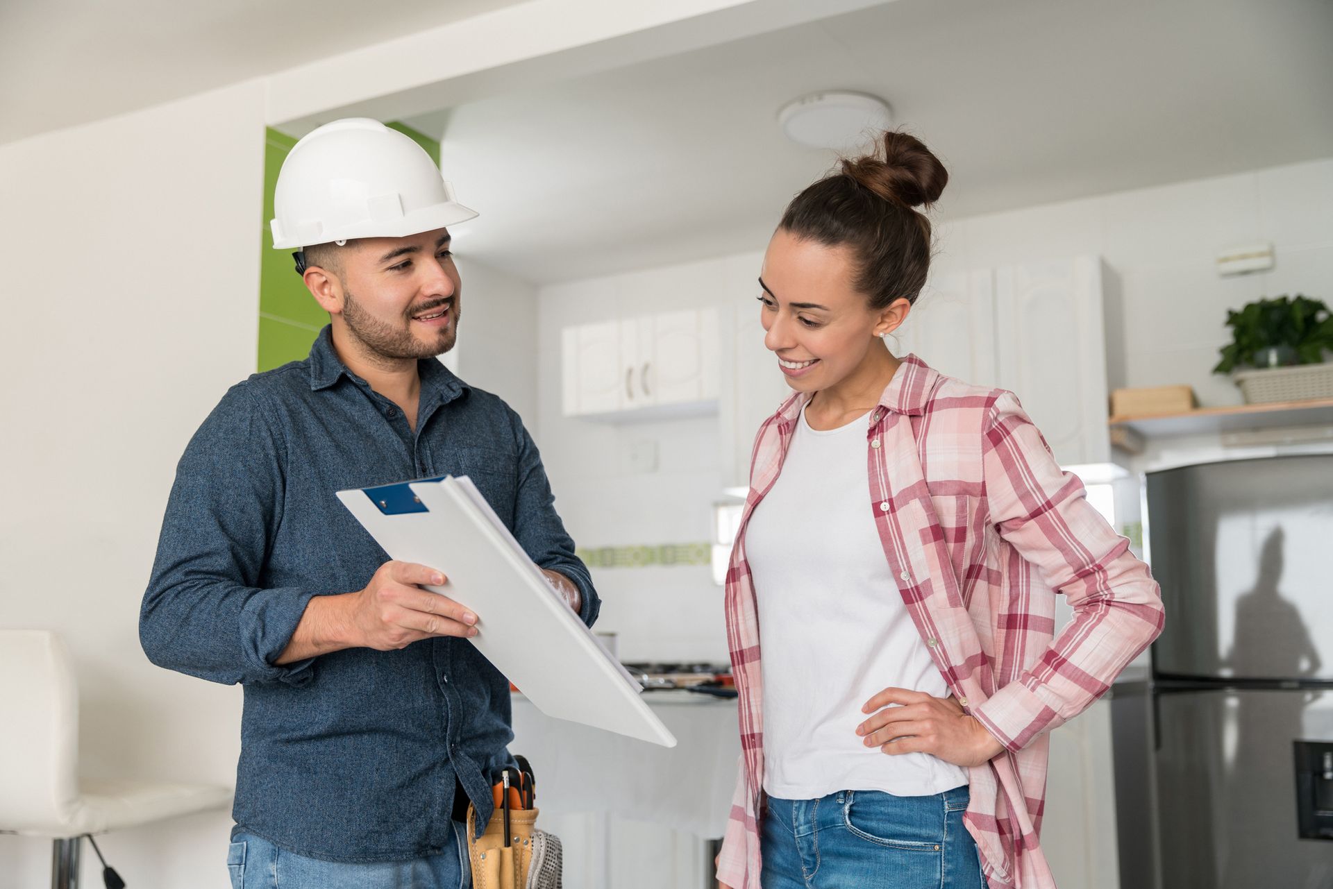 A man in a hard hat is talking to a woman in a kitchen while holding a clipboard.