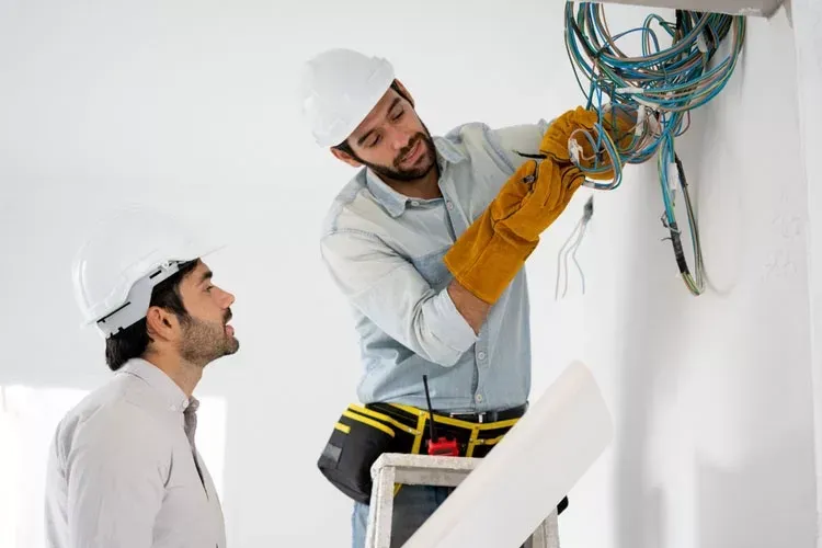 Two men are working on electrical wires on a wall.