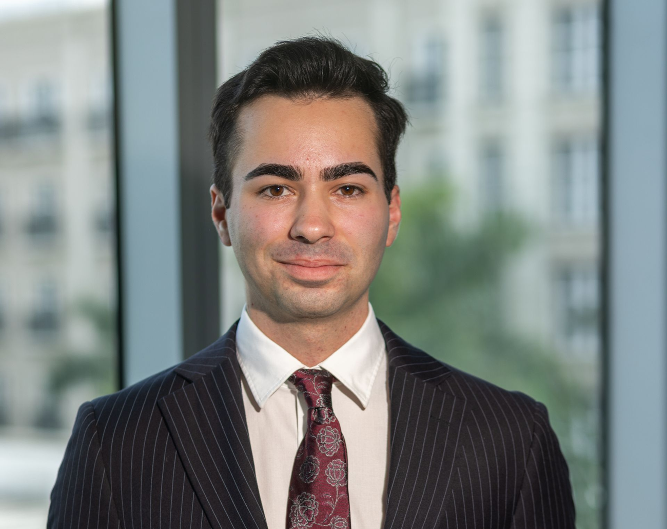 Man in pinstripe suit and tie, smiling, indoors near a window.