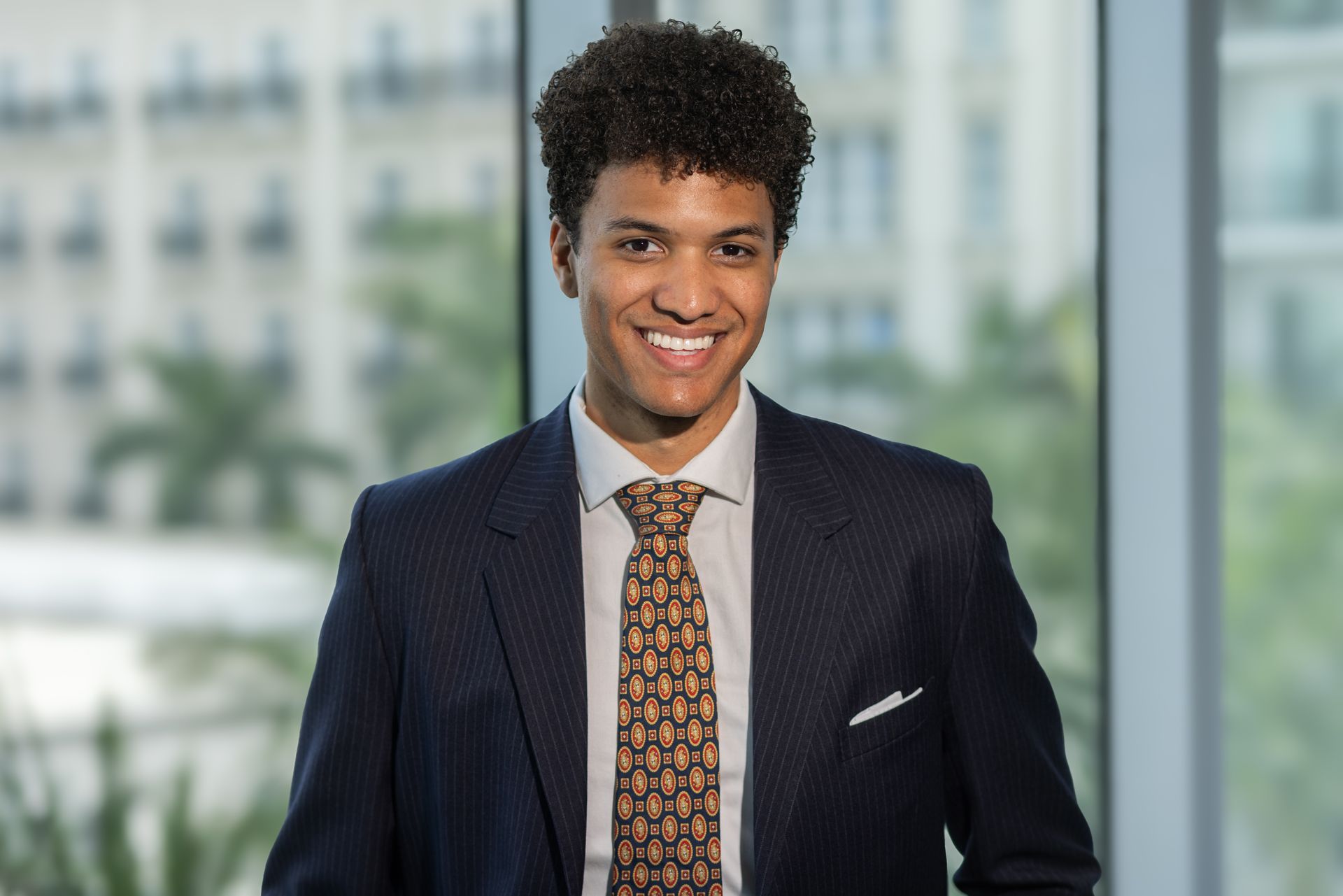 Smiling man in navy suit and patterned tie, standing near a window with a city view.