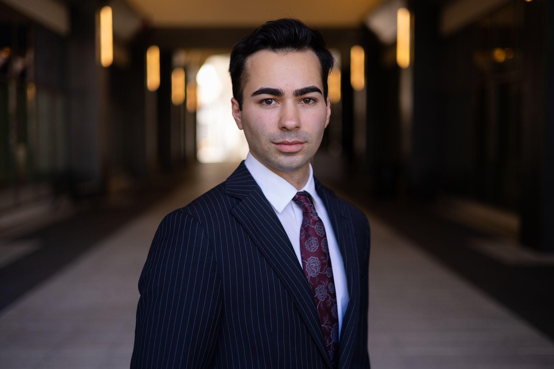 Man in pinstripe suit and tie, looking at the camera, standing in a hallway.