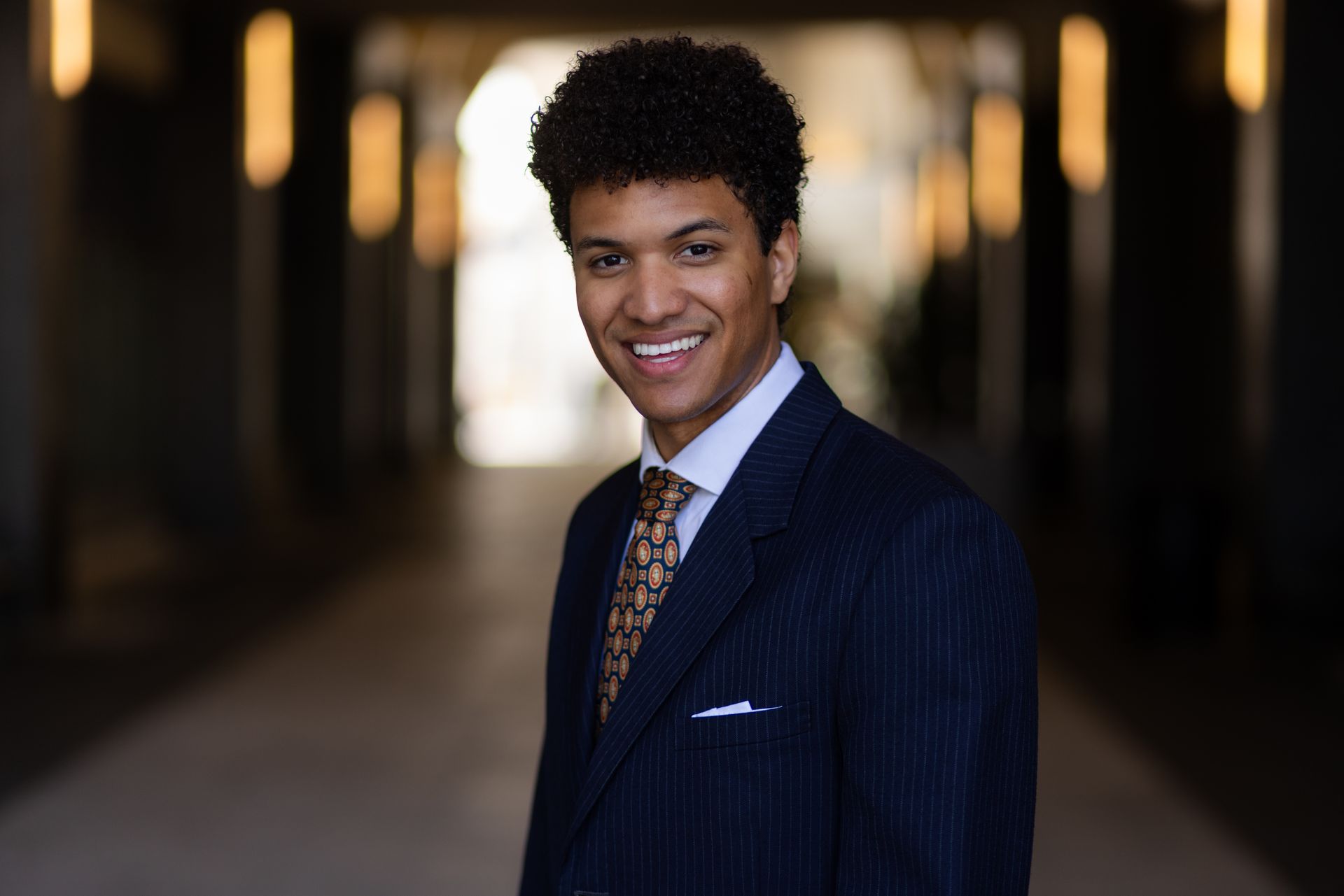 Young man in suit, smiling confidently in hallway with blurred background lights.