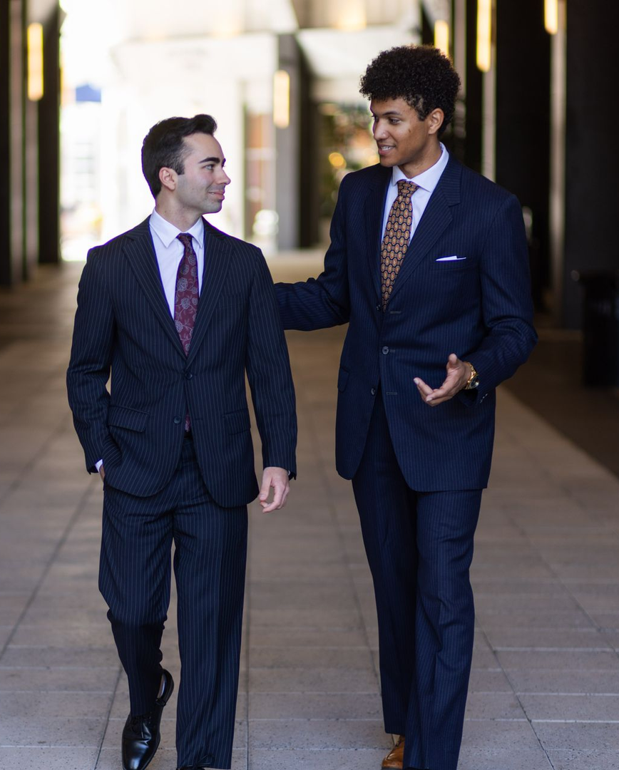 Two men in suits walk and talk outdoors. One has hand on the other’s shoulder. Both wear ties and look at each other.