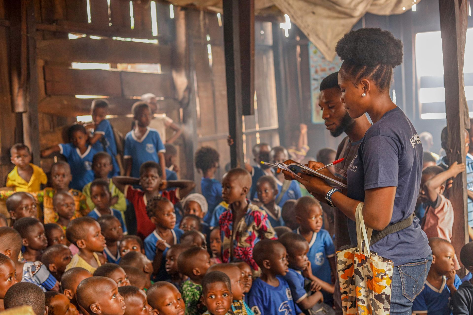 Adults with children in a crowded room, likely a school. Many children in blue uniforms are seated, listening.