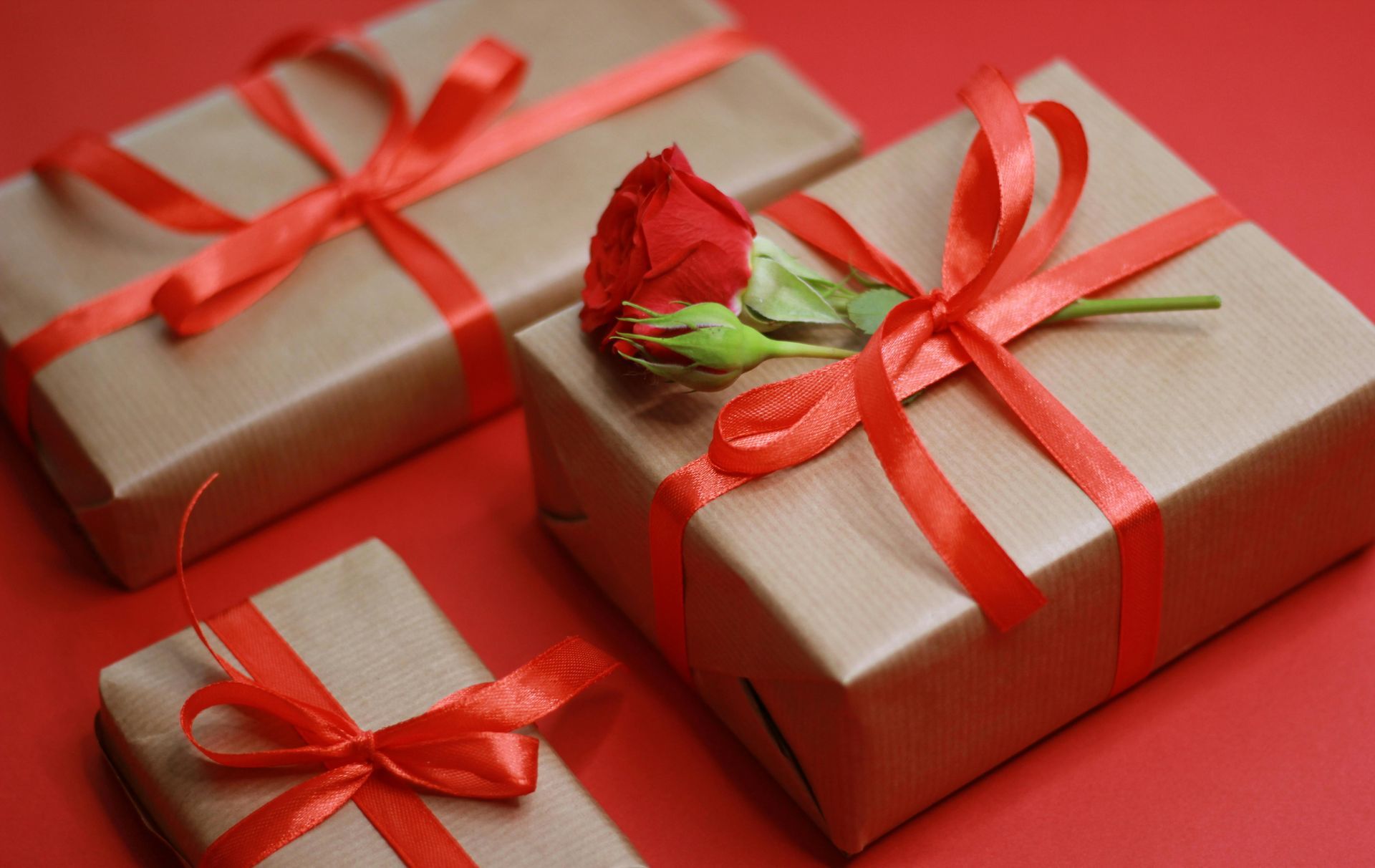 Three brown gift boxes wrapped with red ribbons on a red background, with a small red rose atop the largest box.