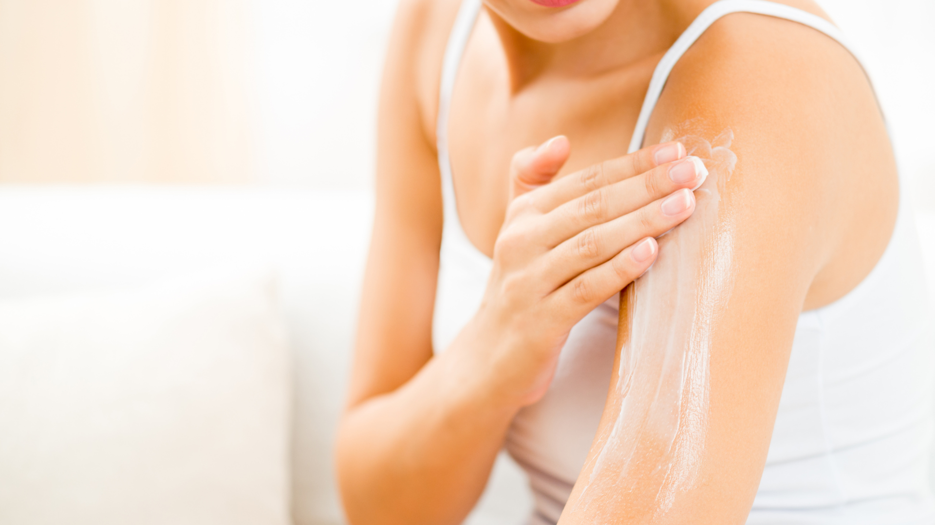 Person applying white lotion to bare shoulder in a bright bathroom