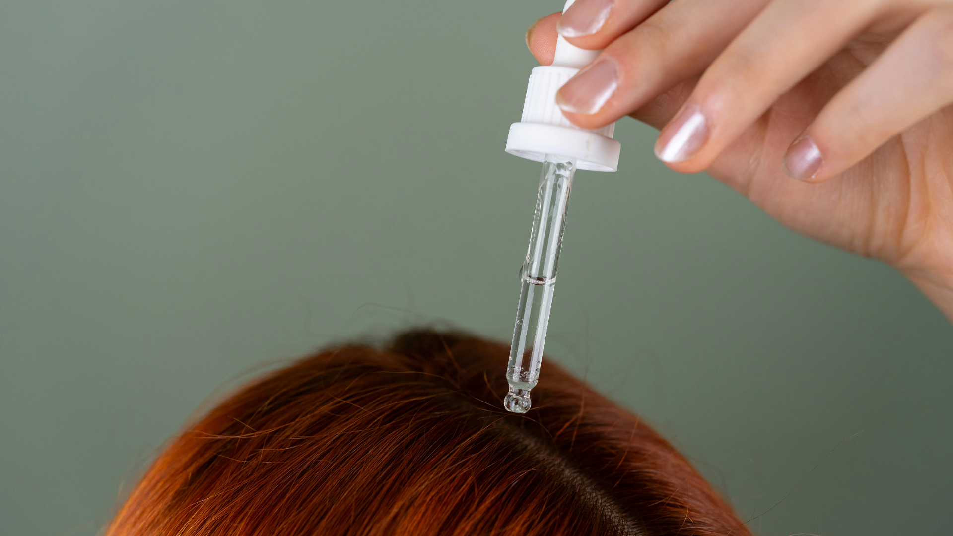Hand holding a dropper over red hair, applying scalp serum