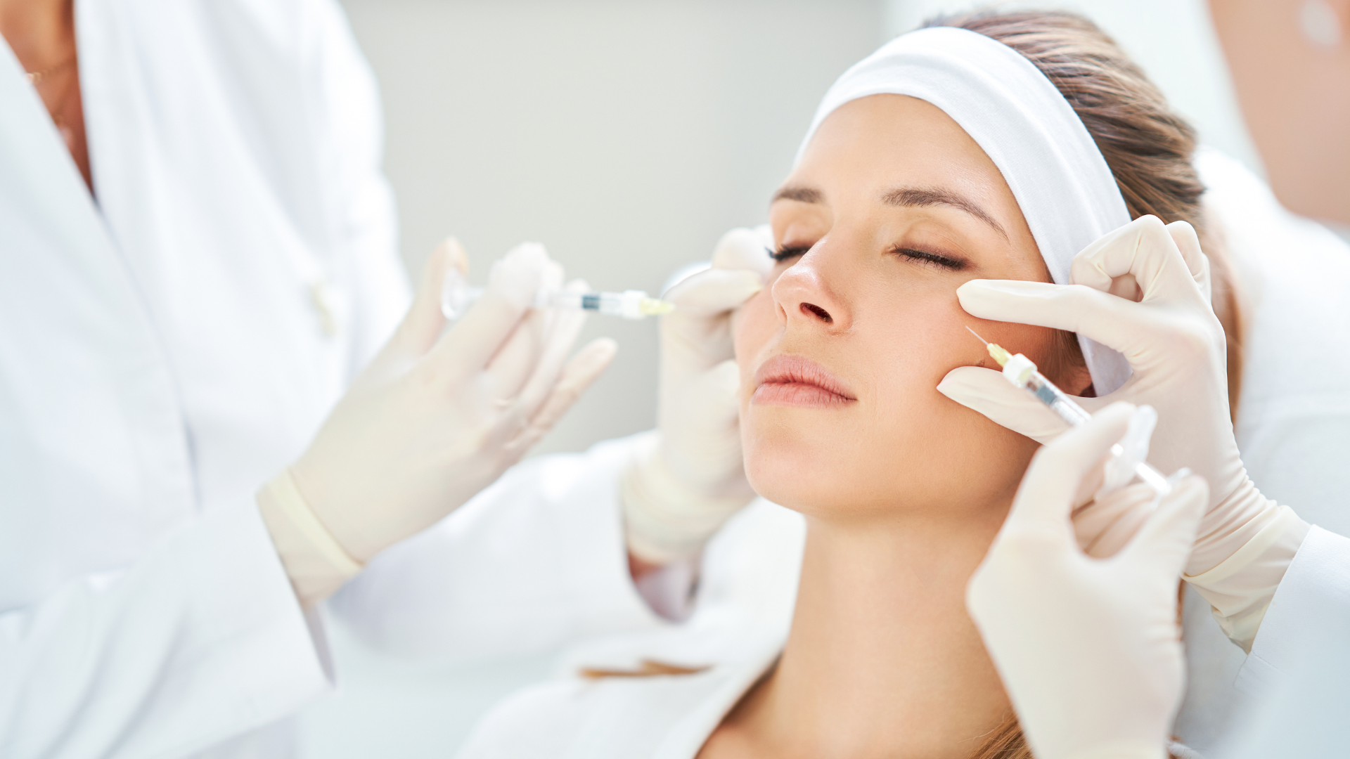Cosmetician applying facial treatment to a woman with white headband in a bright spa setting