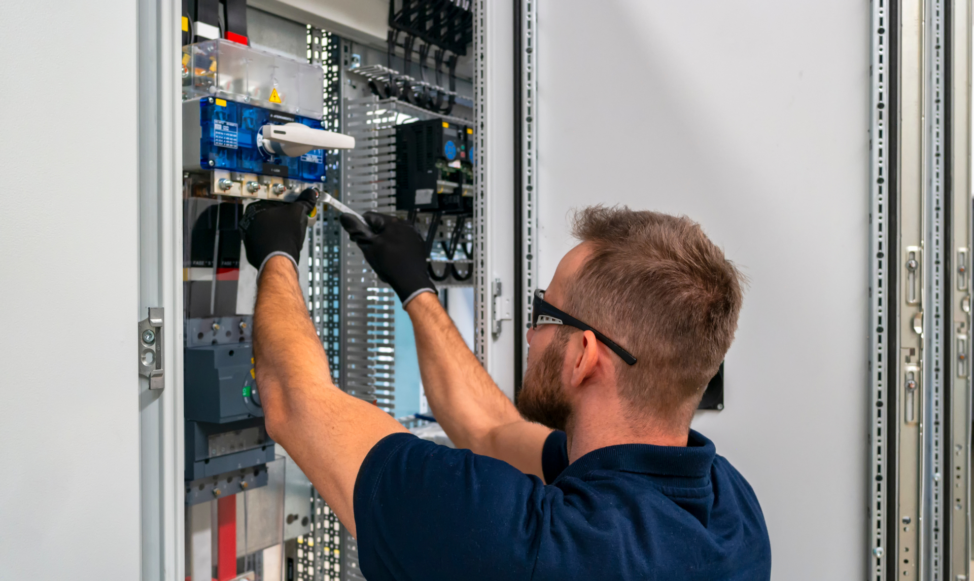 A man wearing gloves and safety glasses working on electrical wiring inside a control panel.