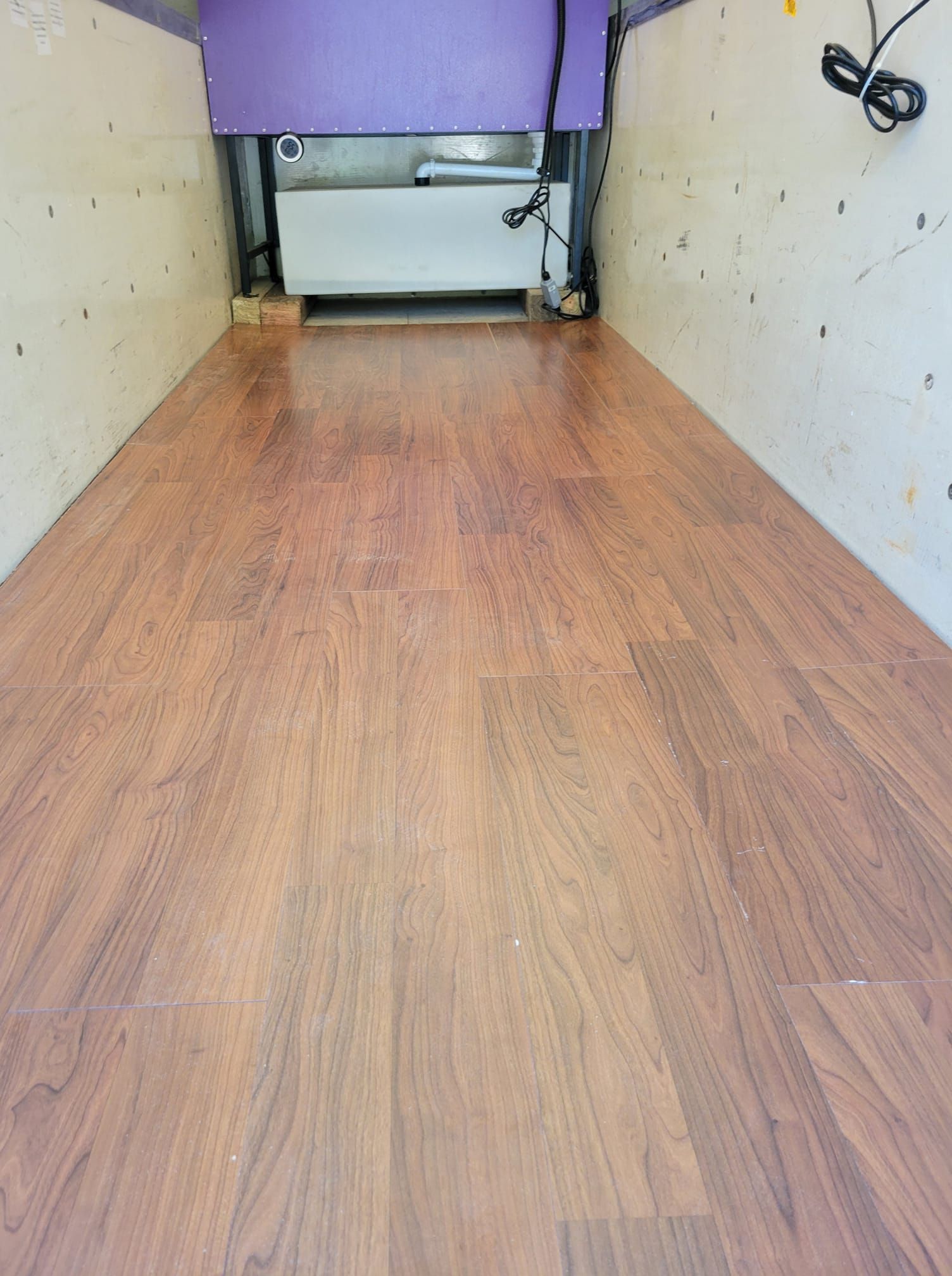 Wooden-look flooring in a rectangular space with light-colored walls. A white rectangular object is in the back.