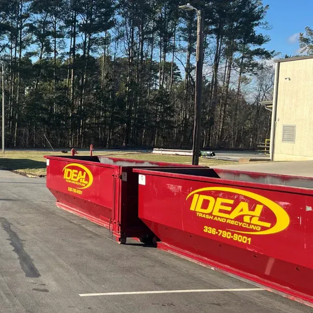 Red IDEAL dumpster on a paved lot beside a beige building and trees