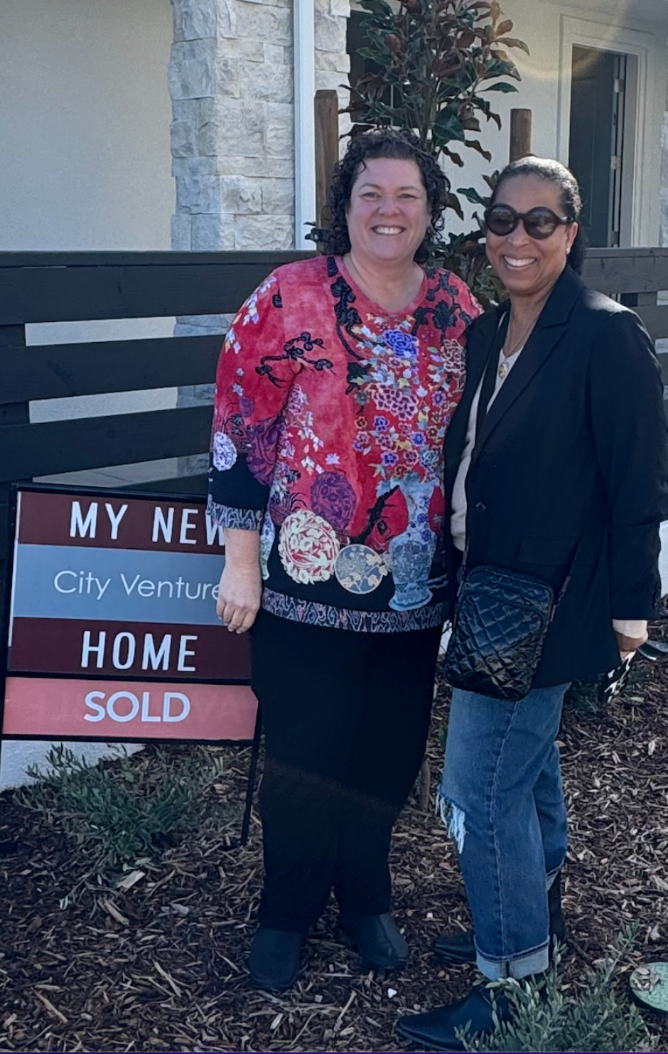 Two women are standing next to each other in front of a sold sign.