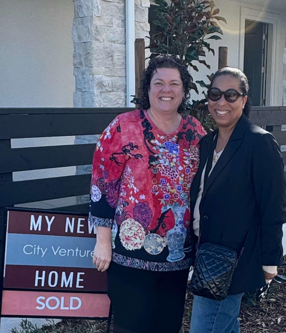 Two women standing next to a sign that says my new city venture home sold