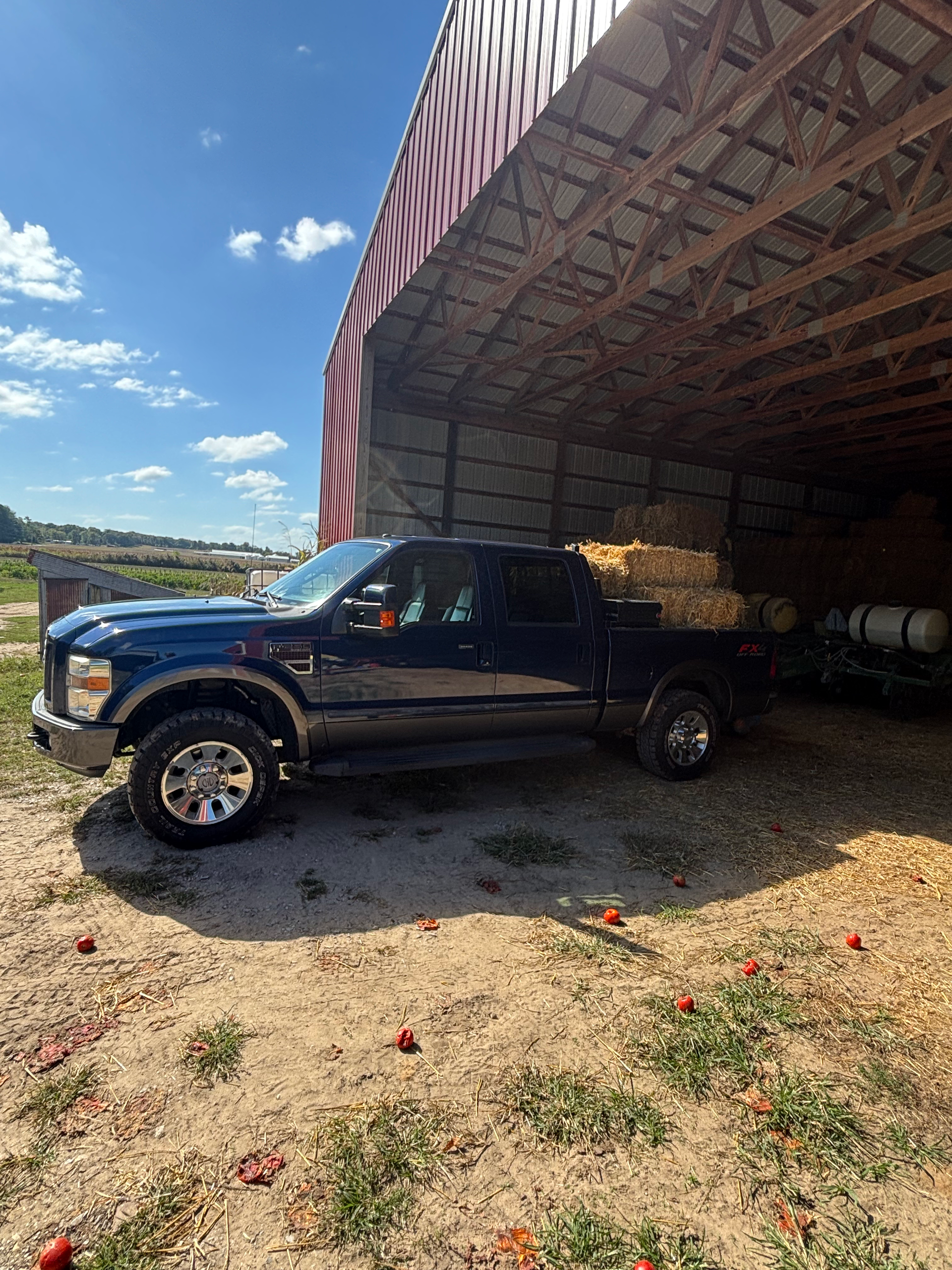 Dark blue pickup truck parked under a barn awning on a sunny day. Hay bales sit in the truck bed.