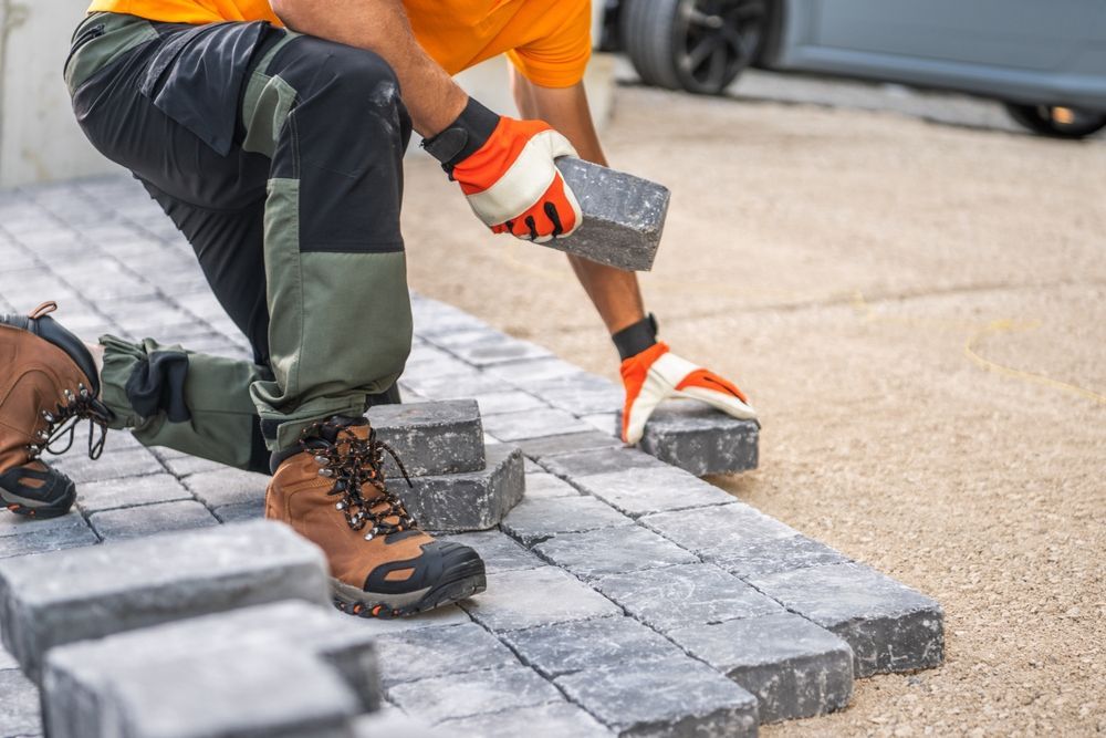 Person laying paving stones, wearing work gloves and boots, outside.