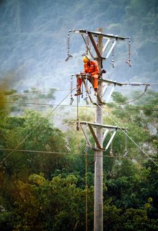 Lineman in orange suit working on a high-voltage power line atop a pole, with forest and hills in the background.