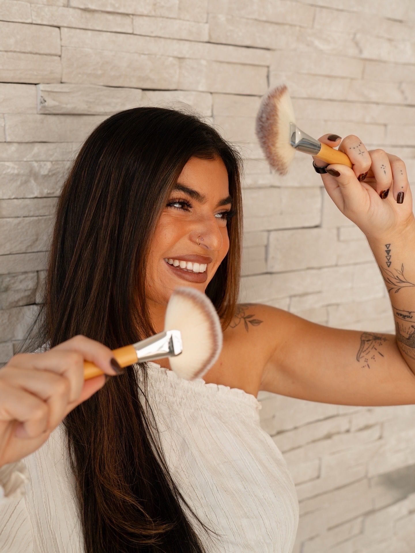 Woman holding facial brushes, smiling in front of a stone wall.