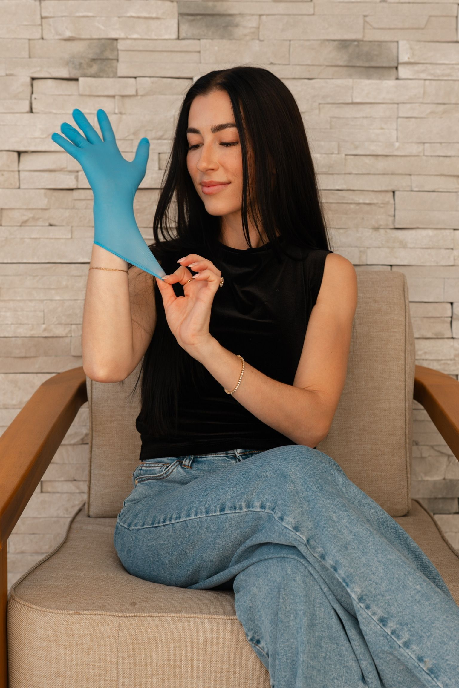 Woman in a black top and jeans putting on a blue glove, sitting in a chair.