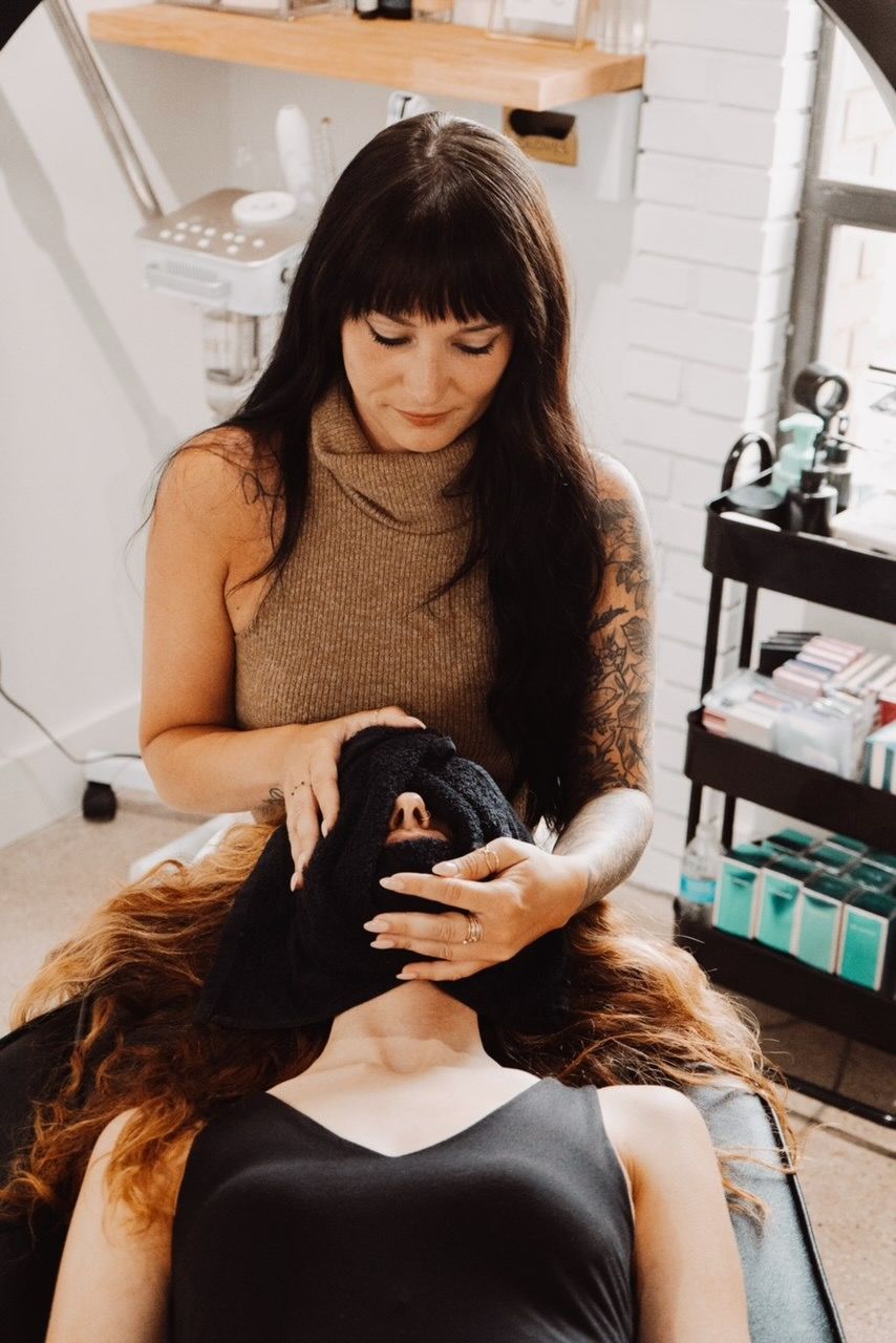 Woman performing facial treatment on another woman. Both are indoors, in a spa.