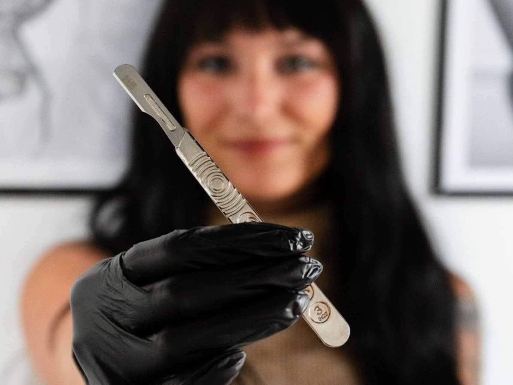Woman in black gloves holds a silver scalpel, smiling.