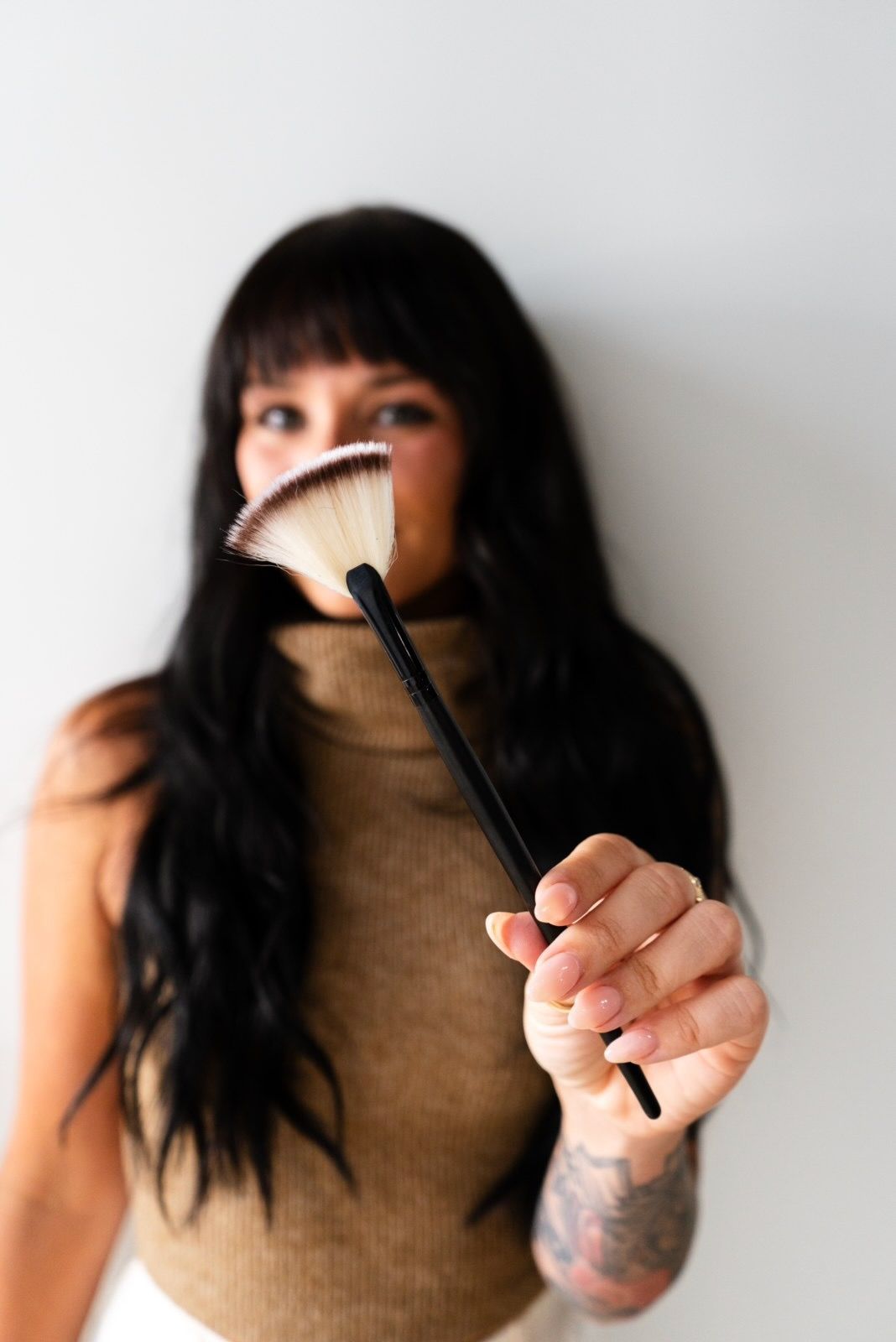 Woman holding a fan-shaped makeup brush in front of her face; wearing a beige top and dark hair.