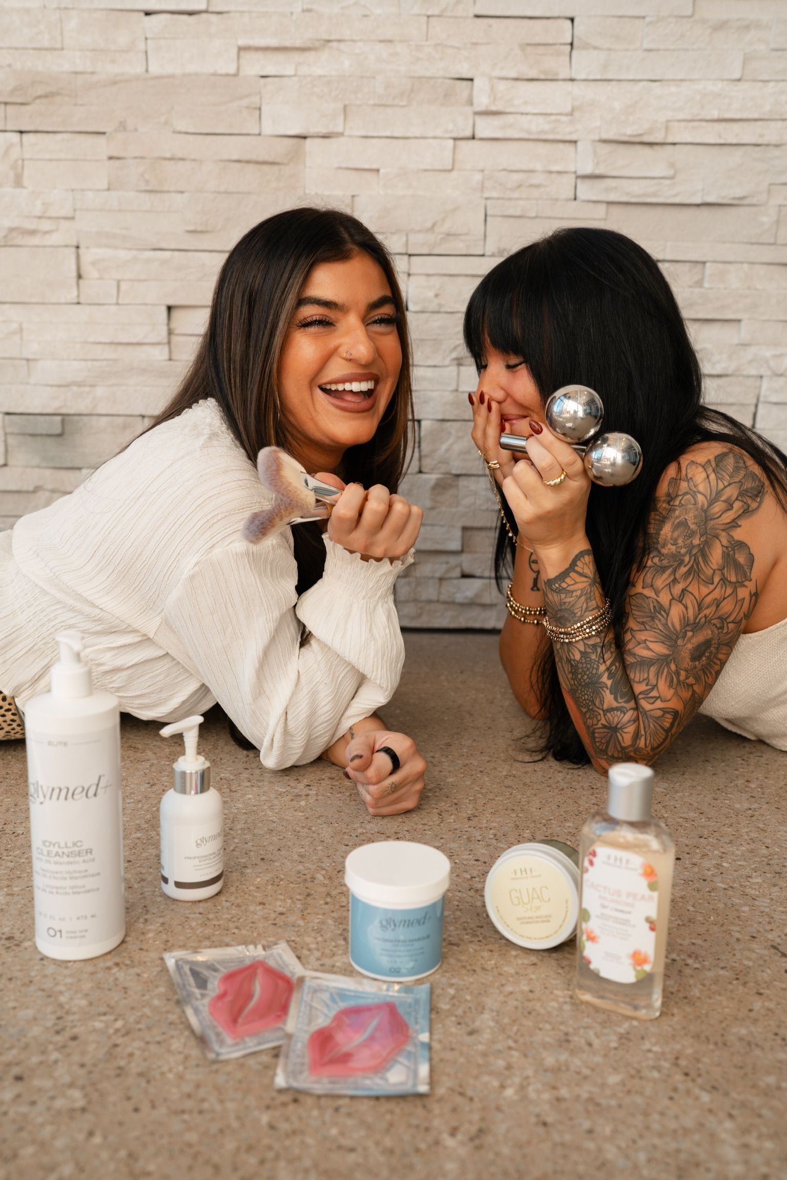 Two smiling women with skincare products. One holds a brush, the other two facial massagers. They're on the floor.