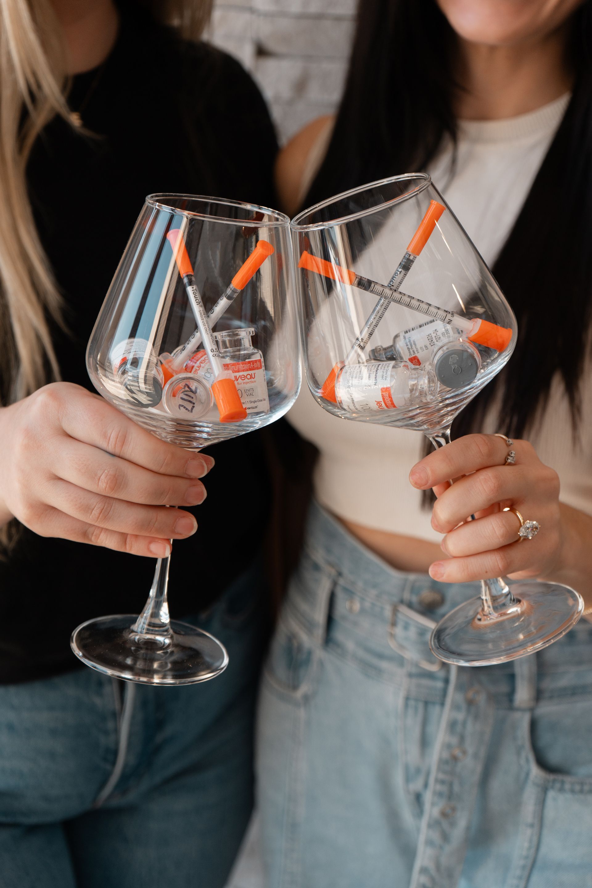 Two people holding wine glasses filled with syringes and vials, clinking them together.