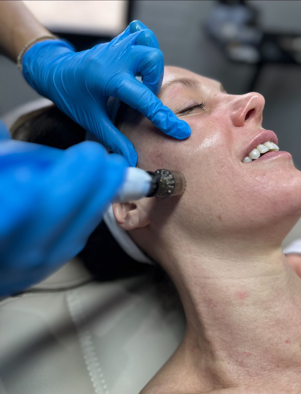 Person receiving a facial treatment. A gloved hand uses a device on her cheek. She has a happy expression.