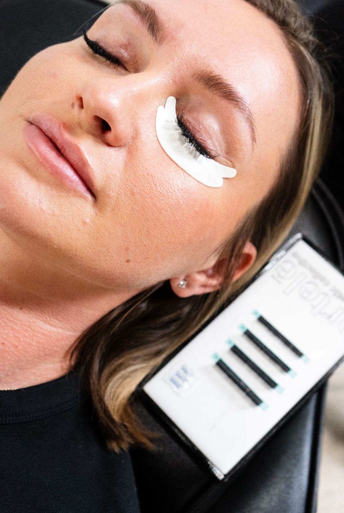 Woman receiving eyelash extensions, eye closed, adhesive pad under eye, lash tray nearby.