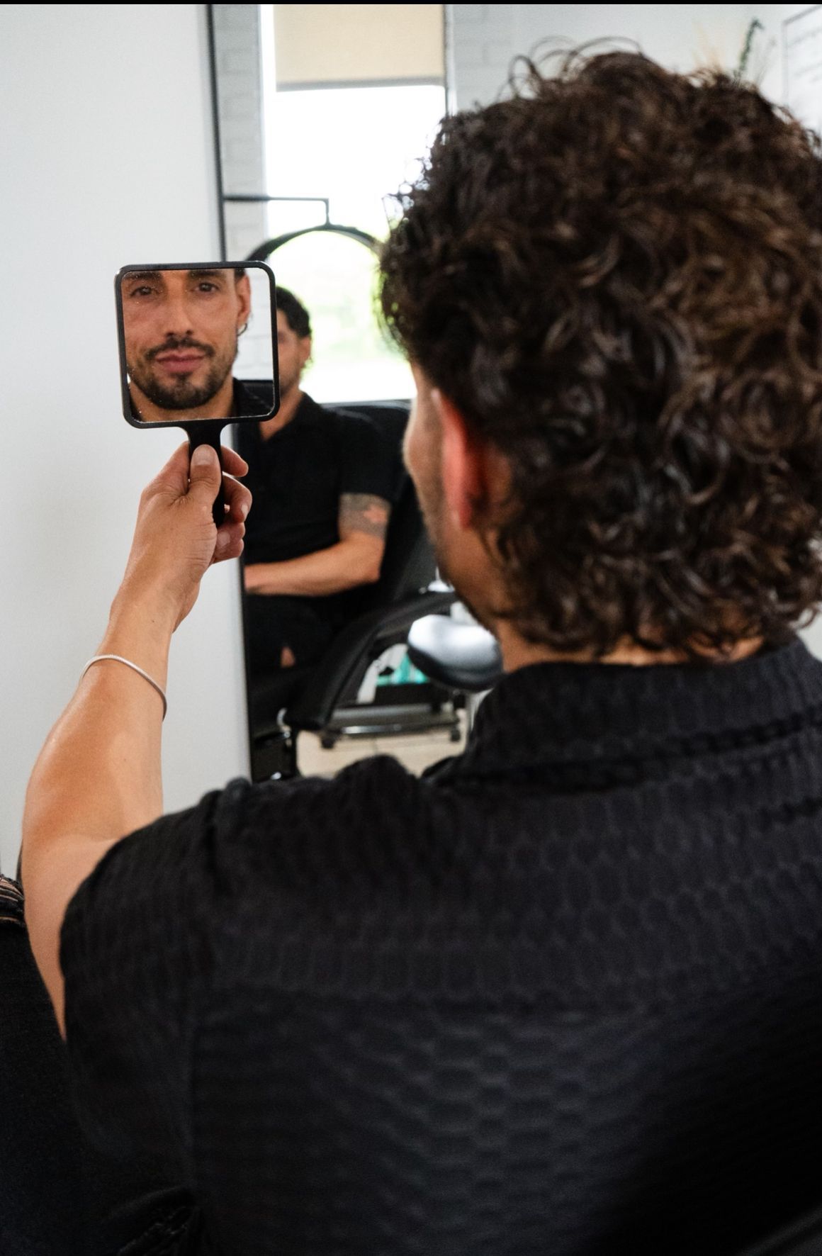 Man looking at himself in a hand mirror in a salon. Black shirt, curly hair, neutral expression.