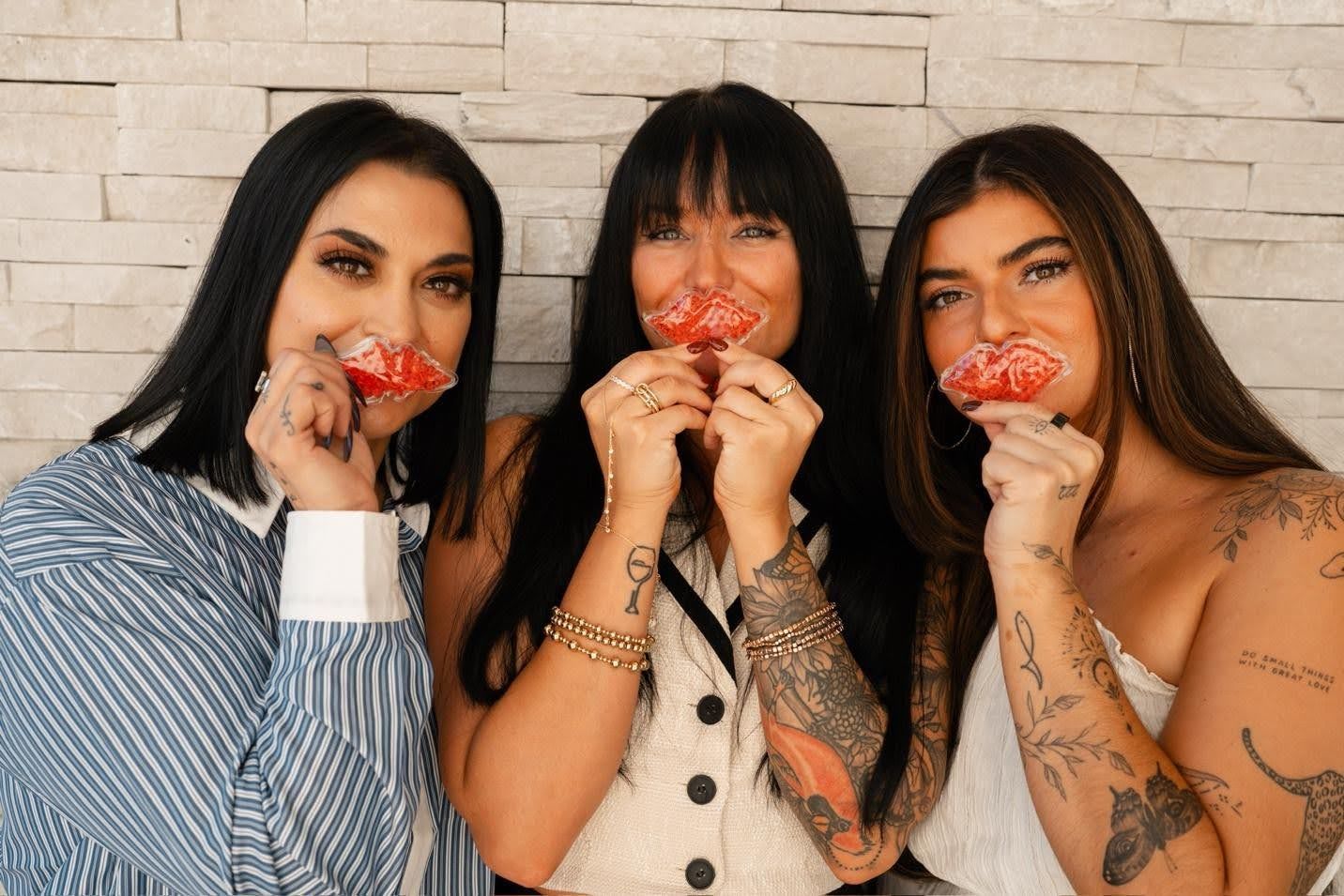 Three women holding lip-shaped lollipops, posing in front of a white brick wall.