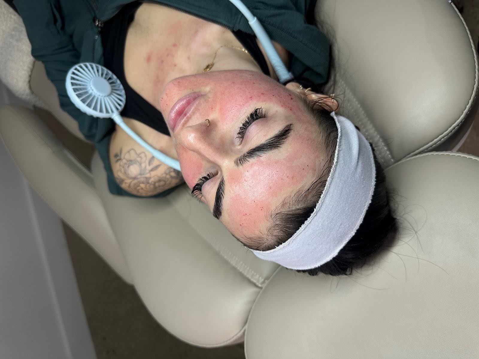Person with pink skin, lying on a chair, with a headband, neck fan, and possibly receiving a facial treatment.
