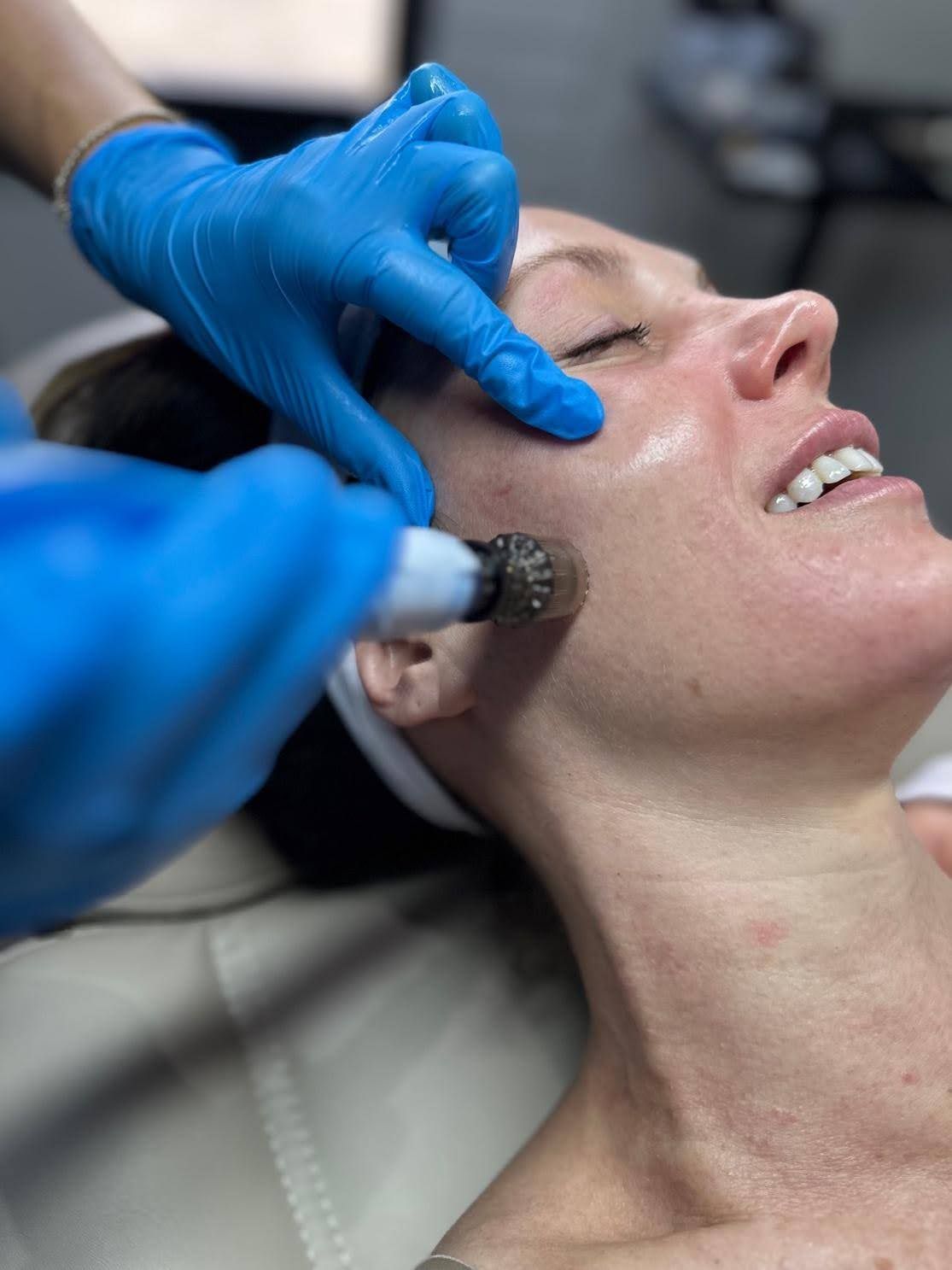 Woman receiving facial treatment; blue-gloved hands holding device to her face, smiling.