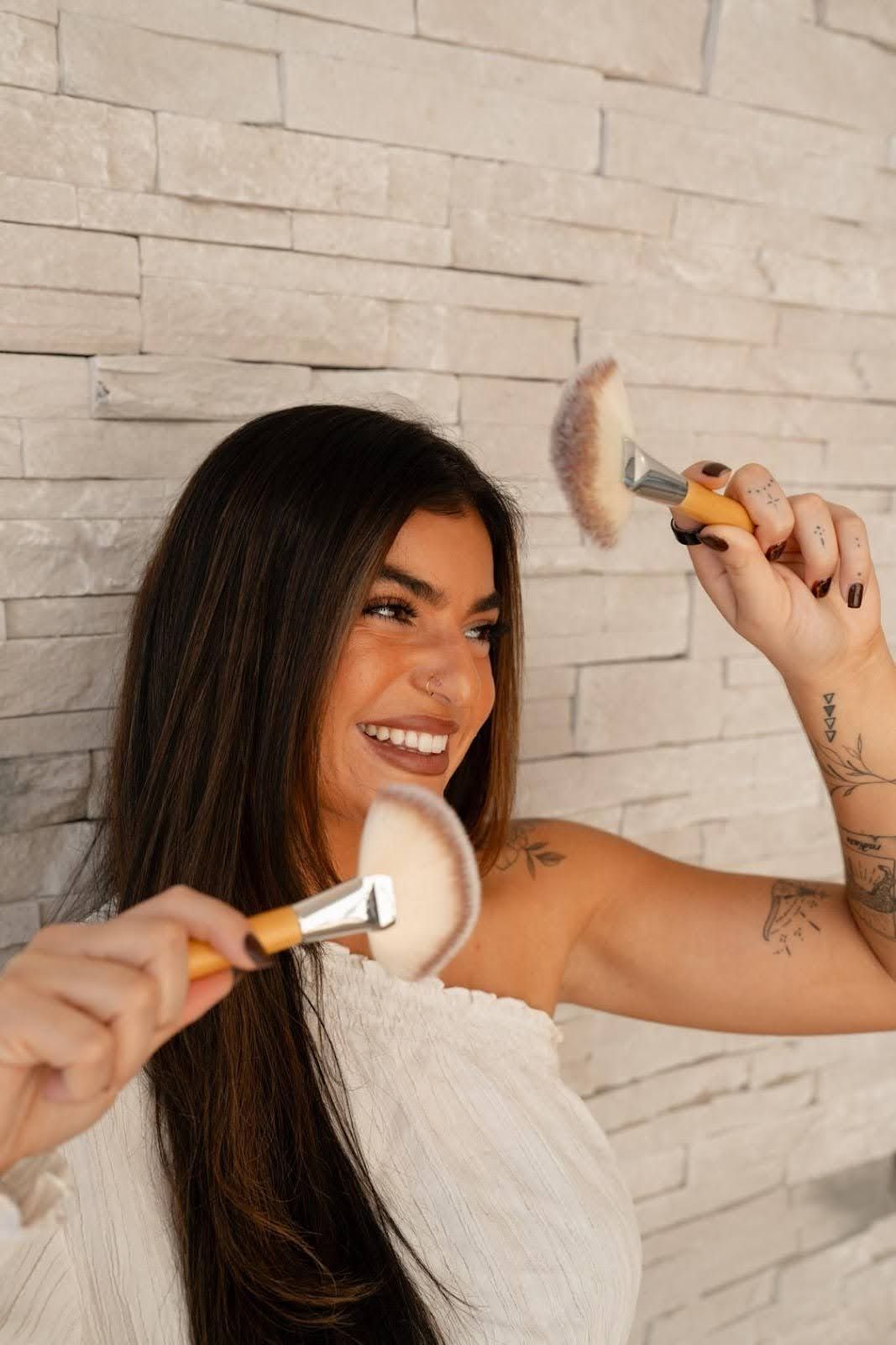 Woman smiling, holding up two makeup brushes. Against a light-colored brick wall.
