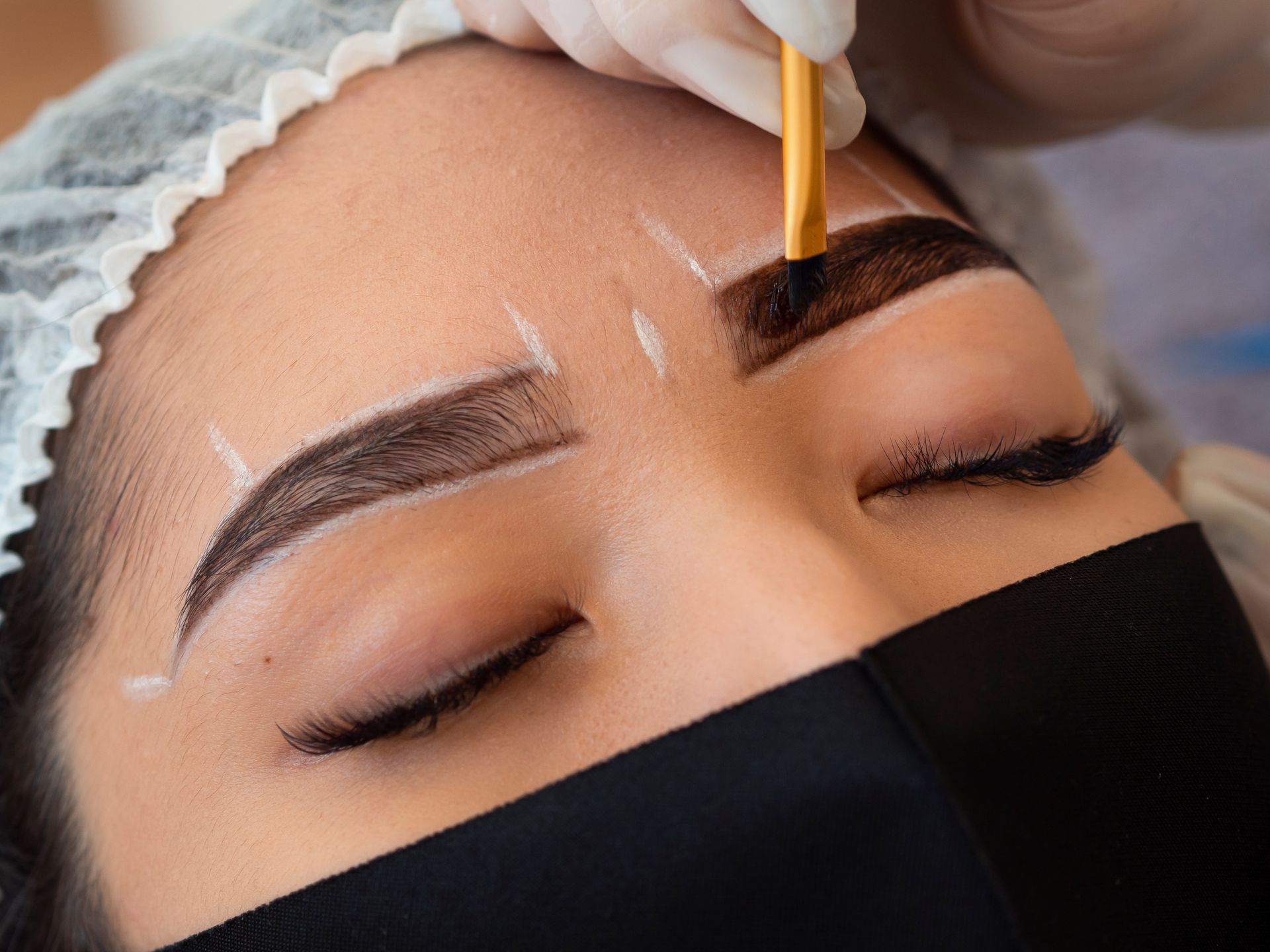 Woman receiving eyebrow tinting. Face mask, white lines mark brow shape, gloved hand applies color.