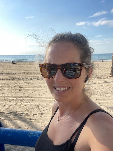 Een vrouw met een zonnebril lacht op een zonnig strand, met golven op de achtergrond.