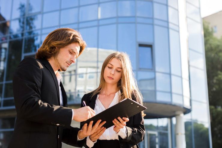 Two business professionals stand in front of a glass building, one signing a document held by the other.